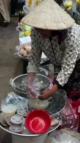 The video shows a bustling outdoor market scene, possibly in Vietnam, with several vendors wearing traditional conical hats. One vendor is shown bagging food items. Other vendors are seen arranging and selling flowers and vegetables. The general atmosphere is busy, with people moving around and engaging in commerce.