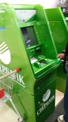 The video shows a person in a black hooded jacket and blue jeans using a green Sberbank ATM. The ATM is located indoors, near a fast-food restaurant with tables and chairs visible through a glass door. A shopping cart is visible near the ATM at the beginning of the video. The person inserts a card into the ATM, then walks toward the glass door. A sign advertising the fast-food restaurant is visible on the glass door, depicting a burger, fries, and a drink. A "Caution Wet Floor" sign is also visible near the entrance. Another ATM, marked "BTБ," is visible to the left of the glass door. The floor is tiled in white, with a dark mat near the entrance.