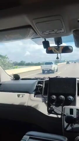 The video shows the interior of a vehicle, looking forward and to the right. The dashboard is visible, along with the side mirror and a portion of the passenger side door. A white pickup truck is driving on a road ahead, on the right side of the frame. The road appears to be a bridge, with water visible to the left. The sky is blue with some clouds.