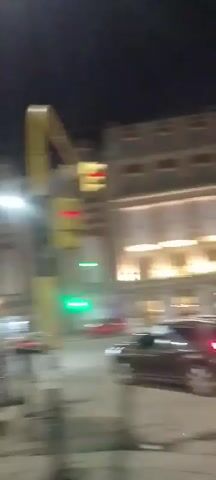 The video shows a nighttime street scene with several cars parked and driving. A tall lamppost with a bright light is visible on the right side of the frame. In the background, there is a multi-story building with lit windows. The street appears to be paved, and there is a sidewalk on the left. There are no clearly discernible people in the foreground. The overall impression is that of an urban environment at night.