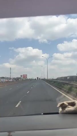 The video shows a view from inside a moving car on a multi-lane highway. The sky is blue with scattered white clouds. On the left side of the road, there's a gas station and billboards. Further down the road, there are a few trucks in the distance. Streetlights line the right side of the highway, with a metal guardrail separating the road from a grassy area. A cloth is visible on the dashboard in the foreground.
