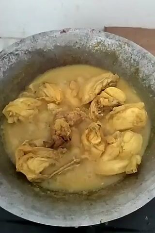 The video shows a weathered-looking aluminum pot sitting on what appears to be a stovetop. Inside the pot is a light yellow, simmering liquid, in which several pieces of cooked chicken are submerged. The chicken has a yellow hue, likely from spices or seasonings used during the cooking process. There's bubbling in the liquid, indicating the food is being heated. The background is a plain, off-white wall.