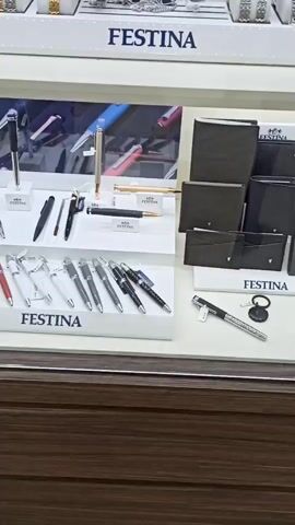The video shows a display case with multiple shelves showcasing Festina brand watches and pens. The top shelf is filled with numerous watches of various styles and colors. Below this, a shelf displays several pens, some with elegant designs, and a selection of men's wallets in dark colors. The bottom shelf also features pens, and further down, a collection of watches similar to the top shelf is visible. The overall scene is well-lit and organized, emphasizing the products on display.