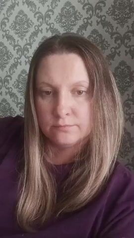 A woman with shoulder-length highlighted hair stands in front of a patterned wallpaper. She wears a purple shirt. The woman begins by looking directly at the camera while she states, “Моя причёска до.” She then uses her right hand to pull her hair behind her ear.