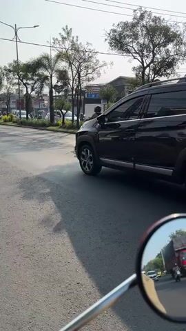 The video captures a rearview shot from a motorcycle looking at a black Mitsubishi Xpander driving on a paved road. The car is moving away from the camera, and its taillights are visible. The road appears to be in an urban or suburban area, with trees and buildings lining the sides. Sunlight is casting shadows on the road, indicating it is daytime. The motorcycle rider's mirror is in the foreground, reflecting a glimpse of other vehicles and the sky. The overall scene is of everyday traffic.