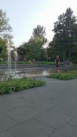 A public park features a large fountain with multiple water jets. In the foreground, a paved walkway is visible with lush green plants bordering the fountain. Several people are present, including a child near the fountain and a person standing on the edge of the water. Other individuals are seen walking in the background and further away within the park. Trees and buildings are visible beyond the fountain area, under a clear sky. The video captures the activity around the fountain and the surrounding park environment.