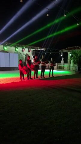 A group of five people are standing on a grassy area at night, illuminated by red and green stage lights. The people are facing the camera. From left to right, the first person is a young male holding a drum. The second person, also a male, is holding a drum. The third, fourth, and fifth people are male and appear to be holding musical instruments. Behind the group is a stage area with a white screen and lights. Above the stage area, multiple beams of white, green, and red light are projected into the night sky. The overall environment appears to be an outdoor event space.