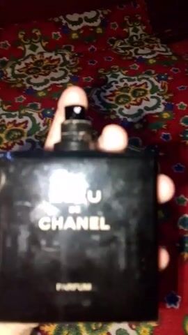 A hand is holding a bottle of Bleu de Chanel cologne over a patterned rug, and sprays the cologne. The rug is red and green with designs of flowers and other patterns on it. The cologne bottle is black with silver text on it, and a black cap. The cologne is sprayed twice. The video is a little blurry and shaky.