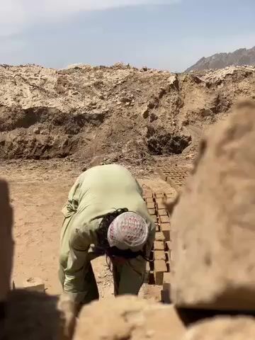 The video shows a man working outdoors in a sunny, dry environment. He is dressed in light-colored, loose-fitting clothing and a white cap. The man is bent over, carefully handling and placing unfired mud bricks on the ground. Several rows of these bricks are already laid out behind him, suggesting they are being dried in the sun. In the background, large mounds of excavated earth are visible, along with distant mountains under a clear blue sky. The ground is sandy and dusty. The man is engaged in manual labor, likely part of a traditional brick-making process in Deh-E Bagh, Afghanistan.