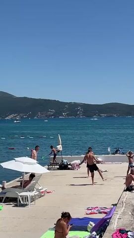 The video shows a sunny day at a beach or waterfront. The foreground features several people relaxing on beach chairs and towels. Two women are visible, one lying on a beach chair under a white umbrella, and another sitting on a towel in the lower part of the frame. In the middle ground, a group of young men are playing a game, possibly beach volleyball or paddleball, on a concrete pier. One person is standing on a platform near a life preserver. The background reveals a calm, blue body of water with a few boats, and a distant shoreline with green hills and buildings. The sky is clear and blue.