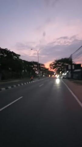 This is a first-person perspective video of a person on a road at dusk. The sky is a blend of purple and orange with clouds. The road is paved and has white and black markings. There are trees and some buildings on the side of the road. Streetlights are on, and there are some vehicles in the distance. The video captures the motion of the person moving forward on the road.