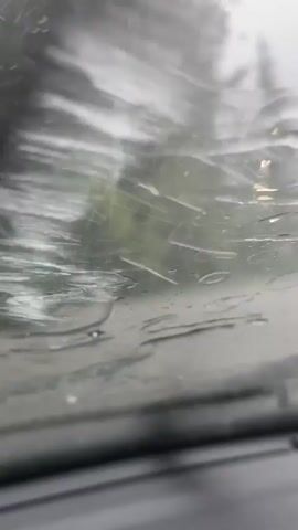 The video shows raindrops hitting a windshield from the perspective of someone inside a vehicle. The background is blurred, showing trees and what appears to be a road. The rain intensifies throughout the short clip, with more and more raindrops appearing on the glass. The windshield wipers are visible at the bottom of the frame.