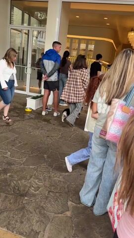 The video shows a crowd of people standing outside a store. Most people are dressed casually, wearing shorts, jeans, t-shirts, and light jackets. Some are carrying shopping bags. The ground is paved with stone tiles. The store has a bright interior with displays visible through the glass windows and doors. There is a small planter with greenery visible on the left. The camera moves slightly, focusing on the back of a woman with long blond hair carrying a large, colorful tote bag.