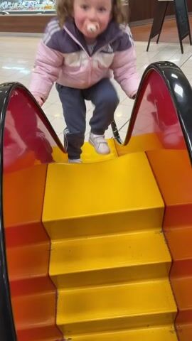 A young child with curly hair and a pacifier is playing on a colorful indoor slide. The child, wearing a pink and purple jacket, grey trousers, and white sneakers, is seen first standing at the top of the slide, looking towards the camera with a wide-eyed expression. They then sit down and slide to the bottom, smiling as they descend. The slide itself is constructed with bright red sides and yellow and orange steps/sliding surfaces.

In the background, there is a brightly lit indoor space, likely a supermarket or a retail area with a food section, given the visible deli-style counter with various items displayed. An adult's legs are partially visible in the background near this counter. The floor is light-colored and tiled. A black A-frame sign with unreadable white text stands further back. The scene suggests a fun, casual moment of play in a public indoor setting, possibly Saarburg, Germany.