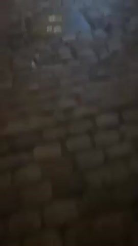 The video shows a night time view of a cobblestone street in what appears to be a European town or city. The camera moves along the street, showcasing several buildings with storefronts. One building is labeled "Bar à Vin" and another says "Brasserie". The buildings are lit from within, and the street is illuminated by streetlights. No people are visible in the video, though one figure is seen moving in the distance. The street is wet in places, reflecting the lights.