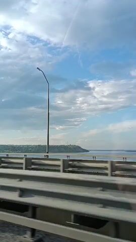 The video shows a view from a vehicle moving across a bridge. In the foreground, there are guardrails of the bridge. In the background, a wide body of water is visible with a distant tree-covered shoreline. The sky is partly cloudy with patches of blue sky. A single street lamp is visible on the right side of the frame.