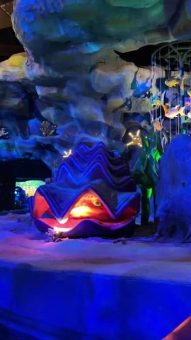 The video shows a brightly colored underwater scene, likely part of a theme park attraction. A large, stylized clam shell with blue and purple outer ridges and a red interior is the central focus. A glowing light emanates from within the shell. A hanging mobile of colorful fish adds to the underwater theme. Starfish and coral decorations are scattered around the sandy base. The entire scene is illuminated with blue light, suggesting an underwater environment.