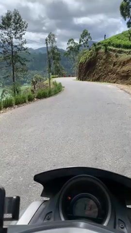The video begins from the perspective of someone riding a scooter. The scooter's dashboard is visible in the foreground, showing a speedometer reading of 79. The road ahead curves to the right. On the left side of the road, there is a steep drop-off with a light blue lake below, surrounded by lush green vegetation and trees. The right side of the road is a steep, grassy embankment. Further in the distance, rolling green hills with scattered trees can be seen under a cloudy sky. The camera pans slightly to the left, offering a wider view of the scenic landscape, including the lake and the hills. The scooter continues on the winding road.