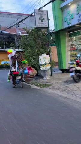 The video shows a blurry, fast-moving scene on a street. In the foreground, a person is riding a bicycle laden with numerous items, including colorful balloons, brooms, and other assorted goods. The bicycle is moving from left to right across the frame. In the background, there is a building with a green facade, likely a pharmacy, indicated by a green cross logo and shelves stocked with products visible through the windows. An older man is seated on another bicycle next to the pharmacy, looking at his phone. The environment appears to be a town or city street, with trees and planters along the sidewalk. The overall impression is of a busy street scene with vendors and daily life unfolding.