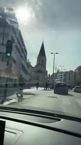 The video, filmed from inside a car, shows a view of a street in a city. The car is moving forward, and the sun is shining brightly in the sky. On the left, there's a modern building, and in the middle, a church with a tall steeple and clock. There are several people walking across a crosswalk in front of the church. To the right, there are more buildings and some flags on poles. Several other cars are visible on the road.