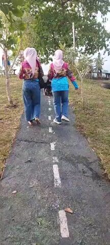 A group of individuals, primarily women wearing hijabs and colorful blouses with blue jeans, are walking away from the camera on a paved path. The path has a dashed white line down the center, resembling a road. The environment appears to be outdoors, with grass and trees on either side of the path. In the background, more trees and a body of water are visible. The lighting suggests it is daytime, possibly late afternoon or early evening, with soft natural light. One individual carries a dark bag over her shoulder. The individuals are walking in the same direction, at a moderate pace. The camera is positioned behind and slightly below the group, capturing their movement away from the viewer.