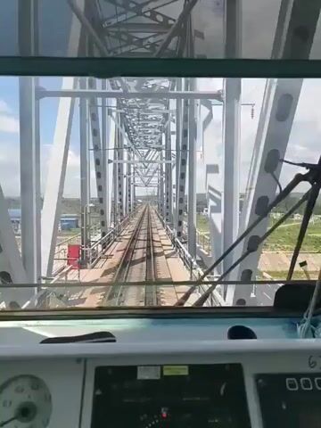 The video provides a first-person perspective from inside a train cabin as it travels across a bridge. The train is moving forward on a track that is part of a large metal truss bridge. The bridge structure is visible overhead and to the sides, constructed from gray steel girders and supports. Beyond the bridge, a wide, open landscape is visible under a blue sky with scattered clouds. The landscape features a railway line stretching into the distance, bordered by fields and some scattered buildings. Power lines and poles run alongside the tracks. The train cabin interior is partially visible in the foreground, with a dashboard displaying various gauges and controls. The camera is positioned looking forward through the windshield.