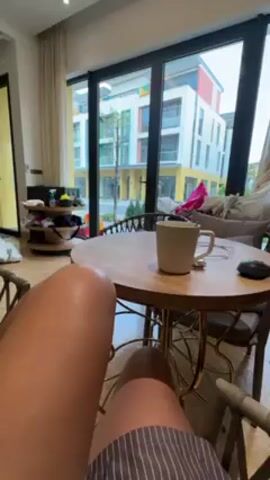 From a first-person perspective, a person wearing gray-striped shorts sits in a wicker chair. A round wooden table in front of the person holds a white mug, a spoon, and a black mouse. Large windows behind the table offer a view of colorful buildings with white, yellow, blue, and red facades. Some greenery and trees are visible outside, as well as what appears to be a stroller. Inside, other furniture and various items are scattered around the room.