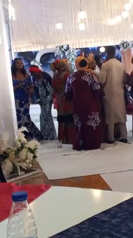 The video shows a group of people dressed in festive attire, possibly at a wedding or celebration. They appear to be dancing or socializing on a white carpet, which is part of a larger, decorated floor.  The room has a white draped ceiling with hanging lights. There's a table in the foreground with a water bottle. The people's movements and interactions suggest a joyful and celebratory atmosphere.