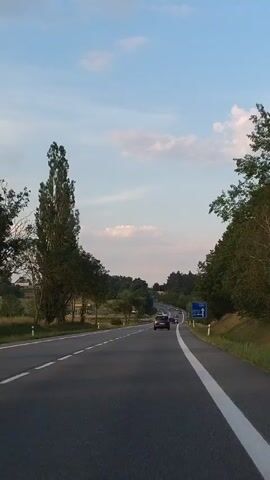 The video shows a view from a moving vehicle on a two-lane road. The road curves to the right and uphill. There are trees and foliage on both sides of the road. In the distance, there are more trees and some buildings. The sky is blue with some clouds. There are several other vehicles on the road, including cars and what appears to be a truck or bus. There is a blue traffic sign on the right side of the road, but the text is illegible.