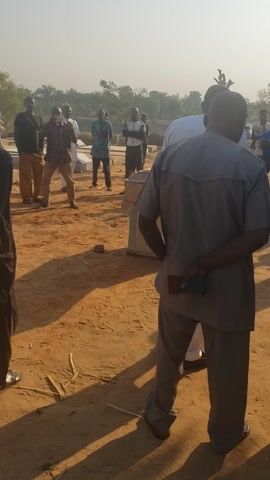 The image captures an outdoor gathering of people in a dry, open area with reddish-brown earth, under bright daylight. The scene, characteristic of Birnawa, Nigeria, suggests a clear and likely warm or hot day, with short, distinct shadows cast by the sun.

In the foreground, a man is prominently featured with his back to the camera, positioned slightly right of center. He wears a grey tunic-style top and matching grey trousers. His hands are clasped behind his back, holding a small green item, possibly a piece of cloth. He appears to be observing or listening attentively.

Further into the scene, numerous other individuals, mostly men, are scattered across the dusty ground. They are dressed in a variety of attire, including white tunics, patterned shirts (one person closer to the camera wears a white and brown patterned long-sleeved shirt), and dark trousers. Their collective stance suggests they are standing and participating in or observing an event. In the background, sparse trees and vegetation line the horizon, beneath a seemingly clear, possibly hazy, sky.

The overall atmosphere appears to be one of solemnity or respectful attention, indicative of a community meeting, religious service, or memorial gathering. There is no visible text within the image itself.