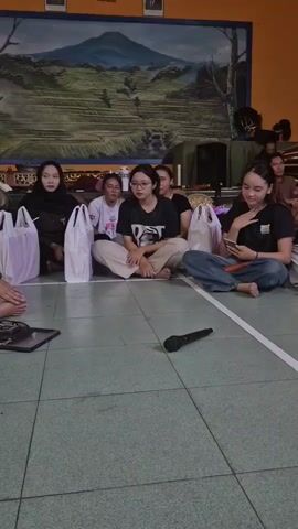 The video shows a group of young people sitting on the floor in a room with a painted mural on the back wall. There are several white bags placed around the individuals. A microphone lies on the floor in the foreground. One of the individuals in the foreground is holding a phone and appears to be looking at it. The lighting suggests an indoor setting, possibly a community hall or school. The people are dressed casually.