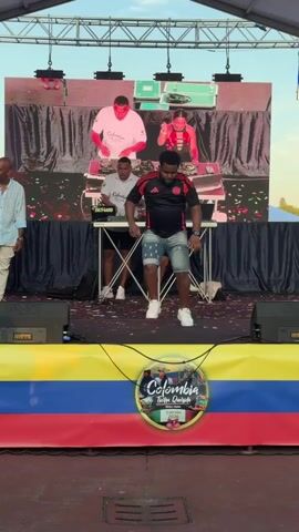 A man in a black t-shirt with red accents and denim shorts dances energetically on a stage in front of a large screen displaying images of DJs. He is wearing white sneakers and appears to be the focus of the performance. Behind him, three other individuals are visible on the stage, two near DJ equipment and one to the side. The stage is decorated with the colors of the Colombian flag. Confetti is scattered on the stage floor. The background appears to be outdoors, with a tent structure overhead.