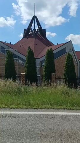 The video shows a view of a church with a modern architectural style. The church has a distinctive roof with a prominent steeple topped with a cross. Scaffolding is visible on the side of the building, indicating that renovations or construction are in progress. In front of the church, there is a row of tall, slender evergreen trees and a black metal fence. A signpost is also visible on the left side of the frame, with directional arrows and place names such as "Warszawa," "Rypin," and "Włocławek." The foreground consists of a grassy area with tall weeds and a paved road, suggesting the viewpoint is from a moving vehicle. The sky is blue with scattered white clouds.