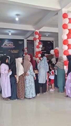 A group of people, predominantly women wearing hijabs, are gathered inside what appears to be a showroom or store. Red and white balloons are strung up, some in vertical columns and others in clusters, decorating the space. In the background, there are posters or banners with logos, one of which appears to be for "YAMAHA" and another for "Amu" with "NMAX TURBO" visible. The people are standing and interacting, some seem to be in conversation or looking towards something specific. The lighting suggests an indoor environment, possibly during the day. One person on the far right is wearing a long, light purple dress, and another to their left is wearing a green hijab and dress. Several individuals are in the foreground, facing away from the camera, or in profile. There's a sense of an event or gathering taking place.