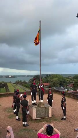 The video captures a ceremonial flag raising event in Galle, Sri Lanka. A group of soldiers in distinctive dark green uniforms with red sashes and caps stand in formation around a flagpole. Two soldiers at the center appear to be in charge of the flag. The Sri Lankan flag, featuring a yellow lion on a maroon background with yellow borders and vertical stripes of green and orange, is visible flying from the pole.

The scene is set outdoors on a raised, dusty area with a stone monument at the base of the flagpole. In the background, a panoramic view unfolds: a body of water, possibly the ocean or a large bay, stretches out, with land visible in the distance. The coastline is lined with trees and buildings, suggesting a coastal town. The sky is overcast and dramatic, with heavy, dark clouds indicating a stormy or at least a very cloudy day, likely late afternoon or early evening given the diffused light.

Several people are present in the foreground, with some filming the ceremony on their phones. One person in a pink top is clearly visible, holding up a phone to record. Another person to the left is partially visible, also holding a phone. The soldiers' posture and the formality of the event suggest a moment of national pride or remembrance. The overall atmosphere appears solemn and respectful. The monument at the base of the flagpole has some visible text, but it is too blurry to be read.