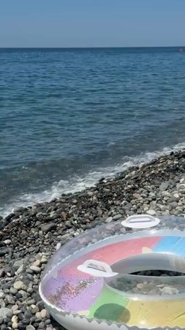 A video shows a close-up of a pebble beach next to the sea on a sunny day. In the foreground, an inflatable swimming ring is visible, partially obscured by the stones. The ring is colorful, with segments of pink, purple, and green, and has glitter embedded within it. The sea is a calm, blue expanse, with gentle waves lapping at the shore. The horizon is clear, meeting a bright blue sky. The camera pans slightly, revealing more of the beach and the water.