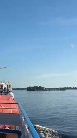 The video shows a view from a boat moving on a body of water. The boat appears to be a ferry or a larger passenger vessel, indicated by the presence of orange life vests on deck and a white railing. The water is calm with ripples on the surface, and the sky is clear blue. In the distance, there is a small island with trees. As the boat moves, the scenery changes, revealing a white tower with a conical roof on a shore, surrounded by trees. Further along the shore, there are houses and buildings, including some red-roofed structures that resemble barns, and a cluster of apartment buildings. A small boat is visible on the water at one point. The overall impression is a journey along a waterway with coastal views of islands and settlements.