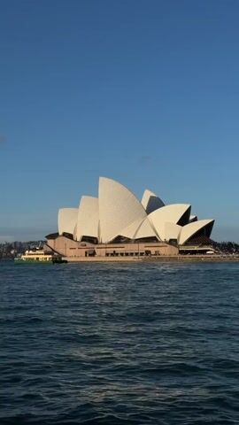 The video shows a slow pan across the Sydney Opera House from the water. The opera house is a multi-venue performing arts center. Its distinctive sail-like shell is white and white. The structure is on the harbor, and the water is a deep blue. The sky is clear and blue. The sun is shining. A ferry is visible in the distance on the left.