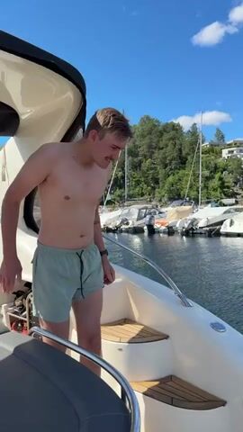 A young man in light green swim trunks jumps from a white boat into a marina on a sunny day. He is shirtless and has short, light brown hair and a mustache. The boat has a black bimini top and chrome railings. In the background, there are several sailboats and yachts docked, with houses and trees on a hillside behind them. The water is a deep blue and calm. The sky is clear with a few white clouds. The man is shown mid-air as he jumps, then splashing into the water, and finally resurfacing with white foam around him. The overall mood is energetic and summery. The timestamp "00:00" is visible at the beginning of the video.