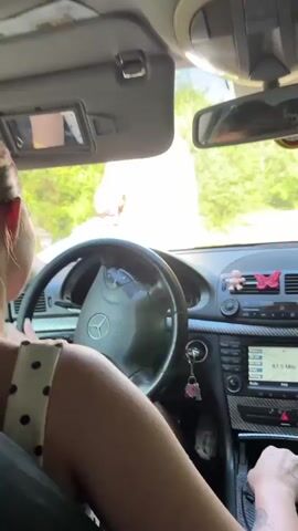 This video shows the interior of a car with two people. A person is driving, with their hands on the steering wheel and gear shift. Another person is standing outside the car, leaning in towards the driver. They appear to be talking or interacting. The car's interior includes a Mercedes-Benz steering wheel, a radio displaying "87.5 MHz," and decorative butterflies on the dashboard. Outside, a rural or natural setting is visible with trees and greenery, suggesting a sunny day. The overall mood seems casual and conversational. The visible text is "87.5 MHz" on the car's radio.