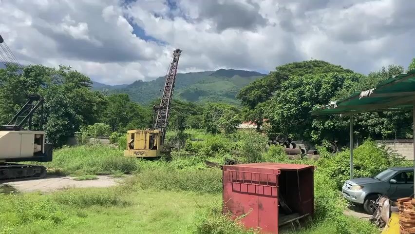This video shows a landscape scene with two large construction cranes. One crane is white and yellow and on the left side of the frame. The second crane is yellow and taller, located in the middle of the frame. Both cranes appear to be stationary and in a field with overgrown grass. In the foreground, there is a red metal container. In the background, there are trees and mountains under a cloudy sky. A silver vehicle is partially visible on the right side of the frame, parked under a lean-to structure. The scene appears to be an outdoor storage or junkyard for heavy machinery.