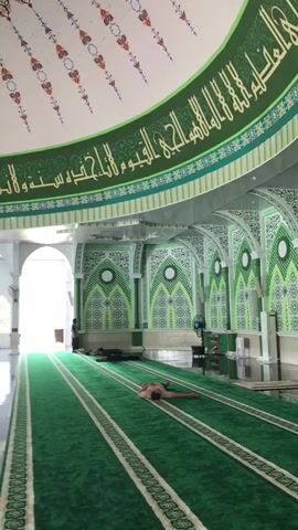 The camera pans inside a mosque. The interior is decorated in green and white, with Arabic script along the curved ceiling. The floor is covered in a thick green carpet with white stripes denoting prayer rows. There is a wooden minbar (pulpit) on the right side of the frame. In the foreground, a man is lying on the carpet. Further away, two other people are also lying down. One person stands near the open doorway in the background. Electric fans stand throughout the room. The room appears empty and quiet.