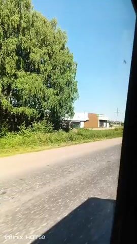 The video captures a view from inside a vehicle traveling on a paved road. On the left side of the road, there is a treeline with a large, leafy tree. Further down the road, a light-colored building with brown and white sections is visible. To the right of the building, a green sign with three circles is present. A silver car is seen driving on the road in the same direction as the camera. A white sedan then overtakes the silver car. The sky is clear and blue. The road appears to be in a rural or semi-rural area.