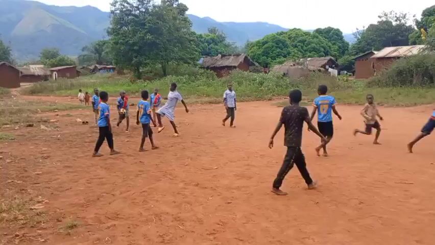 The video shows a group of about a dozen children playing a game on a dirt field in what appears to be a rural village.  Some of the children are wearing matching blue and orange jerseys. There are houses in the background, some of which appear to be traditional huts, and a mountain is visible in the distance. The children are running around, kicking what is likely a ball, although it’s out of frame.  They are playing on a large, open dirt field. Some children are standing around watching the game. One child walks into frame on the right side.