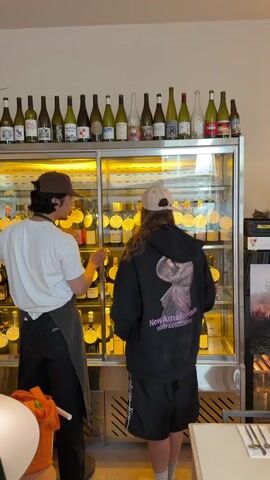 Two people are standing in front of a glass-front wine refrigerator. The person on the left is wearing a white t-shirt, black pants, a brown hat, and an apron. They are gesturing towards the wine refrigerator with their left hand. The person on the right is wearing a black hoodie with an image on the back, black shorts, white socks, and a tan baseball cap. They are looking at the wines in the refrigerator. Behind them is a table with silverware on it. Above the refrigerator are a row of wine bottles. The room appears to be in a restaurant.