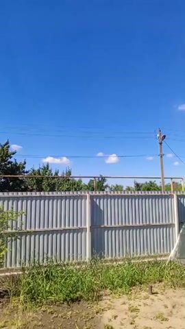A video captures a sunny day outdoors. A tall, gray corrugated metal fence dominates the foreground, with lush green grass and dirt at its base. Beyond the fence, a line of trees with dark green foliage is visible against a vibrant blue sky dotted with a few white clouds. Utility lines are strung across the sky, and a utility pole with a fixture on top is present on the right side of the frame. The sun shines brightly, casting shadows and illuminating the scene. The camera appears to be stationary, with no discernable movement.