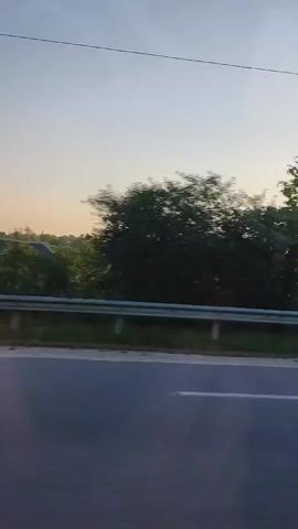 This video captures a car's journey through a rural landscape in Chetroșica Veche, Moldova, during the late afternoon or early evening. The sun, low in the sky, casts a warm glow, creating lens flare and illuminating the scene. The time of day is suggested by the soft light and the long shadows.

The view from the car's window shows a road with white lane markings, indicating it's a paved road, likely a highway or a major rural route. Alongside the road, there are trees with lush green foliage, some of which are quite tall. In the distance, hints of a residential area or farm can be seen, along with structures that appear to be wind turbines. The sky is a clear, pale blue, with some wispy clouds.

The activity is a car traveling, with the camera capturing the passing scenery. The motion blur of the car in the foreground and the trees in the background reinforces the sense of movement. There are no people clearly visible. The overall mood is peaceful and serene, typical of a scenic drive through the countryside as the day draws to a close. The weather appears to be fair. No visible text is present in the video.