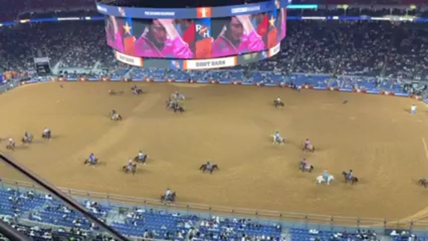 The video shows a rodeo arena with several riders on horseback practicing their skills. The arena is filled with dirt, and the riders are spread out across the space. Overhead, a large screen displays an image of a young man, possibly a competitor. The arena is surrounded by seating, and the stands are filled with spectators.