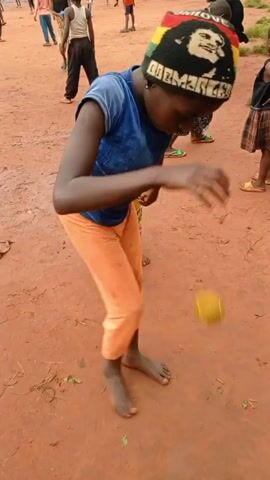 A young person with dark skin is barefoot and wearing a blue t-shirt, orange pants, and a colorful hat with a portrait on it. They are standing on a reddish-brown dirt field and kicking a small, yellow fruit-like object with their bare foot. Other children are scattered around in the background, some standing and watching, others walking or playing. The setting appears to be a village or rural area with simple houses in the distance, and a cloudy or misty mountain backdrop. The video seems to capture a casual, playful scene among a group of children outdoors.