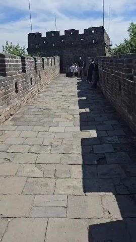A paved walkway of the Great Wall of China with people walking. The wall is made of grey bricks and has battlements along the top. The sky is blue with some clouds. In the background, there is a watchtower. A group of people are gathered near the watchtower, some taking pictures. Two men are walking away from the watchtower and towards the camera. The man in the foreground is wearing a black shirt and pants, and the man behind him is wearing a white shirt and shorts. They appear to be tourists.